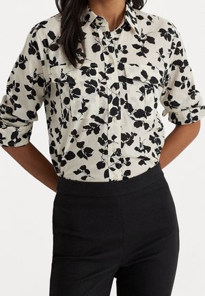 Vrouw draagt een wit hemd met knopen en zwarte bloemenprint en opgerolde mouwen, gecombineerd met een zwarte broek met hoge taille.