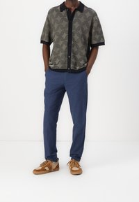Mann in kariertem kurzärmligem Hemd, blauen Hosen und braunen Sneakers, der mit den Händen in den Taschen vor einem weißen Hintergrund steht.