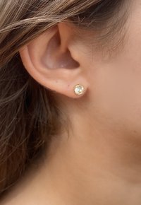 Orecchini a bottone in oro con una pietra preziosa trasparente in un montaggio circolare, posizionati nell'orecchio, che mettono in mostra un metallo liscio e una pietra brillante.