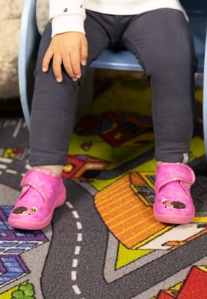 Peuter zittend op een blauwe stoel, gekleed in donkere legging en roze pantoffels met egel- en bijenmotieven op een kleurrijk speelkleed.
