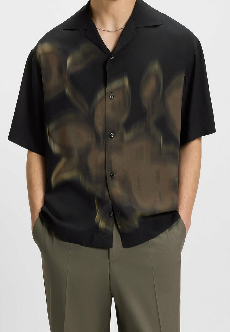 Homme portant une chemise noire à manches courtes boutonnée avec un motif abstrait marron flou et un pantalon vert olive, les mains dans les poches.