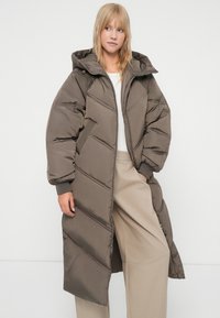 Abrigo largo y acolchado marrón con capucha, con costuras diagonales, abertura lateral y puños acanalados, combinado con pantalones beige.