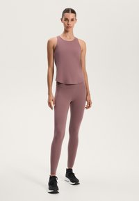 Light mauve athletic tank top with a rounded neckline and draped hem, paired with matching fitted leggings. Black athletic sneakers complete the look.