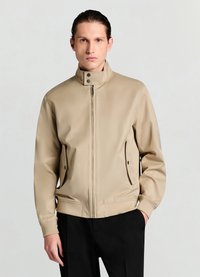 Blouson aviateur beige avec col montant, fermeture éclair à l'avant, boutons-pression et poches latérales. Texture lisse et mate avec coupe ajustée.
