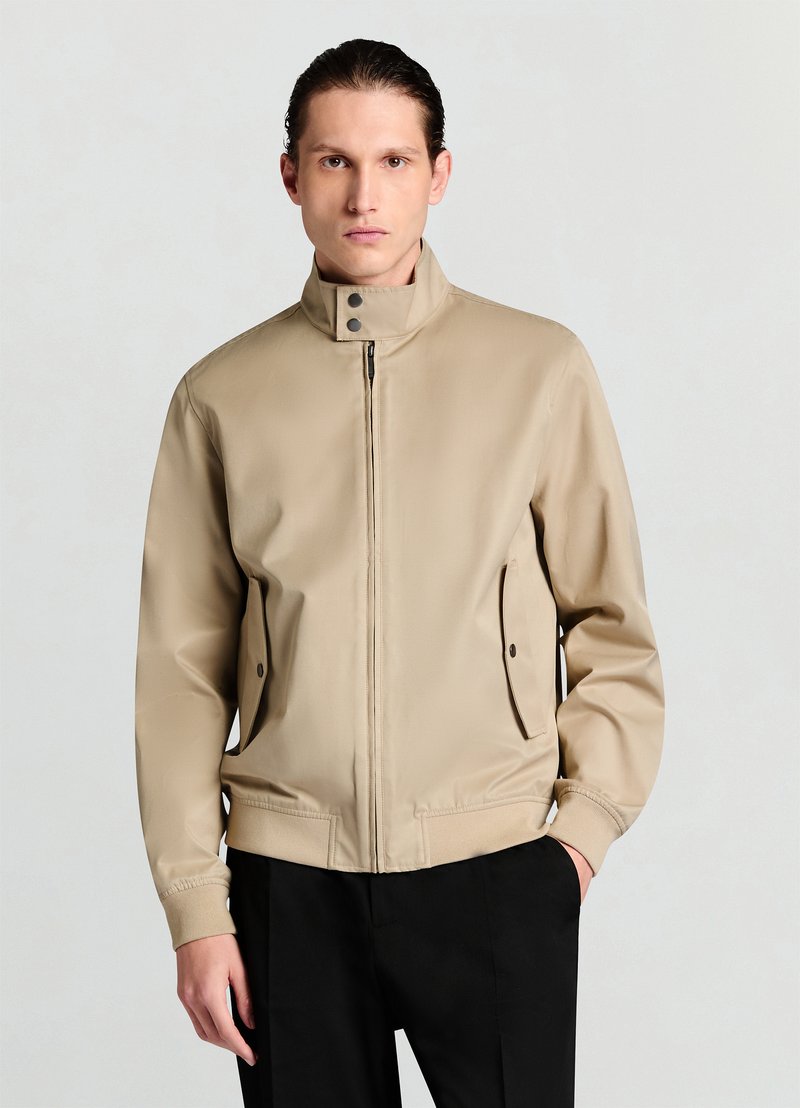 Blouson aviateur beige avec col montant, fermeture éclair à l'avant, boutons-pression et poches latérales. Texture lisse et mate avec coupe ajustée.