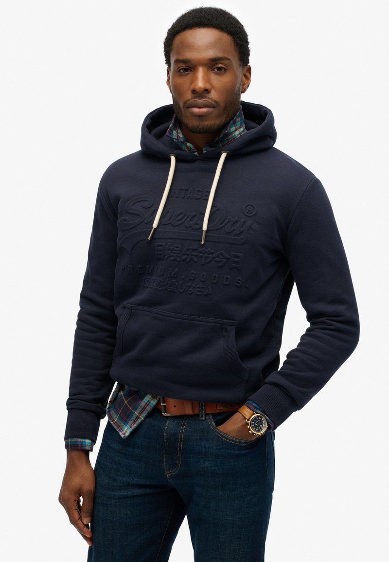 Sudadera navy con bolsillo delantero y capucha con cordón, con logotipo de la marca en relieve. Lleva puesta sobre una camisa de cuadros y combinada con jeans oscuros.
