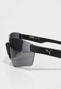 Lunettes de sport noires avec une large lentille grise, présentant une finition mate, un design angulaire et un logo Puma en métal sur le côté.