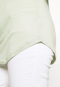 Světle zelený trikový top s krátkým rukávem a zakřiveným lemem, vyrobený z měkké tkaniny. Nosený přes bílé kalhoty, které ukazují hladkou texturu a jednoduchý design.