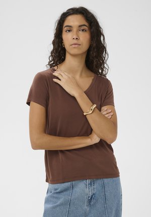 Žena s kudrnatými vlasy, oblečená v hnědém krátkém tričku, světle modrých džínách, zlatém náramku a zlatých kruhových náušnicích, pózující se založenýma rukama.