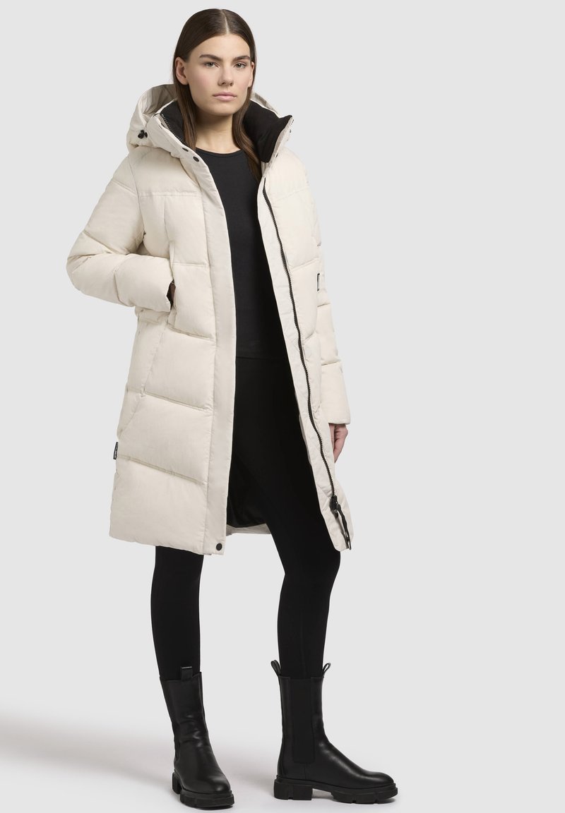 Langbeige lange Daunenjacke mit Kapuze, mit steppdesign, schwarzem Reißverschluss und seitlichen Taschen. Tragen über einem schwarzen Oberteil und Leggings.