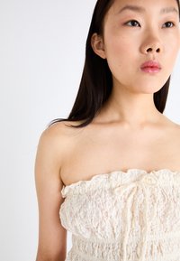Top senza spalline di colore crema con un design strutturato e volants e un dettaglio a nodo sul davanti. Il tessuto presenta motivi floreali e una vestibilità elastica.