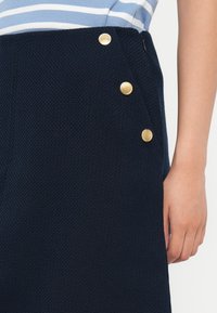 Námornícky textúrovaná sukňa s tromi zlatými gombíkmi na boku, oblečená s svetlo modrým a bielym prúžkovaným topom, ruka položená pri boku.