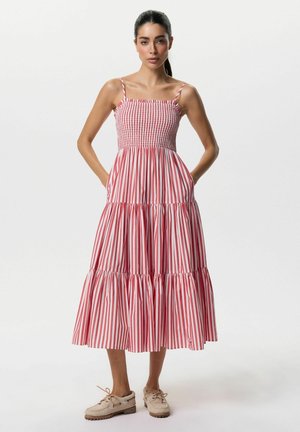 Kobieta stojąca, ubrana w czerwoną i białą, pionowo prążkowaną midi sukienkę bez rękawów z marszczonym biustem i kieszeniami, w połączeniu z beżowymi, sznurowanymi butami.