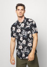 Chemise noire à manches courtes avec des motifs floraux et feuillus en beige et vert. Présente un col et une fermeture à boutons sur le devant.
