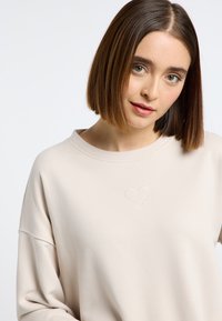 Beige sweatshirt met een relaxed fit, ronde halslijn en geborduurd hartdetail. Gemaakt van zacht materiaal met een gladde textuur.