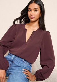 Burgundijas blūze ar V veida kakla izgriezumu, pūkainām piedurknēm un gludu faktūru. Apvienota ar augstas jostas vietas zilu džinsu, radot ikdienišķu apģērba stilu.