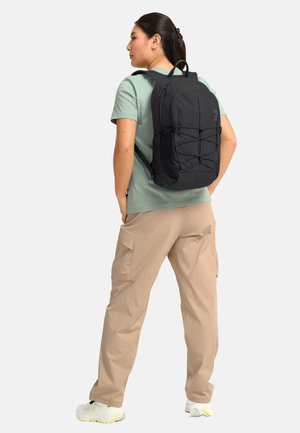 Frau in grünem T-Shirt, beigen Cargohosen und weißen Turnschuhen, die einen schwarzen Rucksack mit elastischem Kordel-Detail auf der Rückseite trägt.