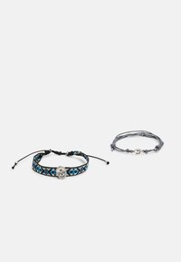 Conjunto de dois pulseiras: uma apresenta um padrão tecido em preto e azul com um pendente de caveira prateada, a outra é um modelo trançado cinza com um símbolo da paz.