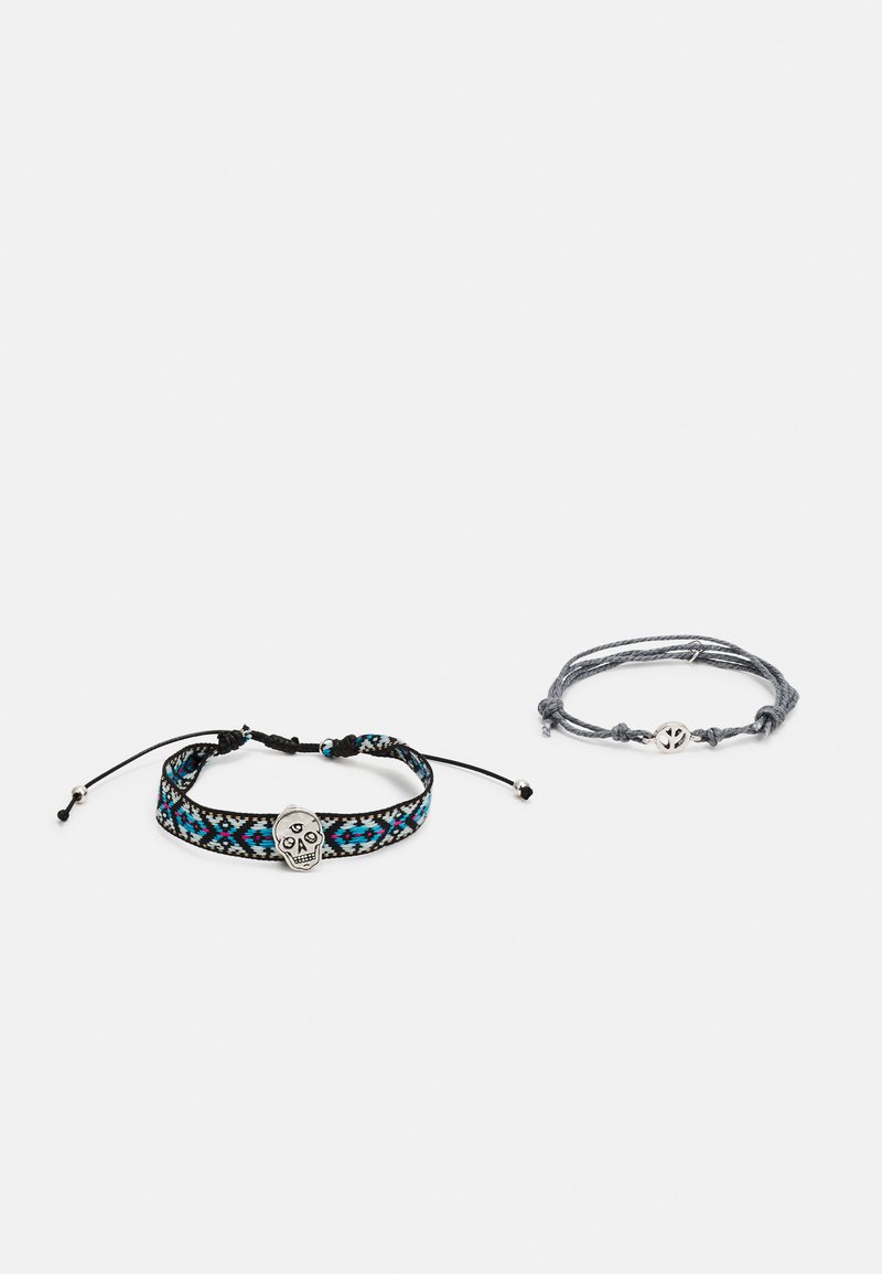 Conjunto de dois pulseiras: uma apresenta um padrão tecido em preto e azul com um pendente de caveira prateada, a outra é um modelo trançado cinza com um símbolo da paz.