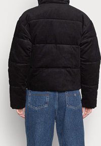 Černá manšestrová prošívaná bunda s hustou prošívanou výplní, vysokým límcem a zaoblenými rameny. Doplněná modrými džíny s kapsami na zadní straně.