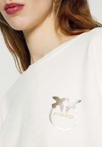 Biała bluza z okrągłym, metalicznym złotym logo z stylizowanymi motylkami oraz napisem „PINKO” umiejscowionym po lewej stronie.