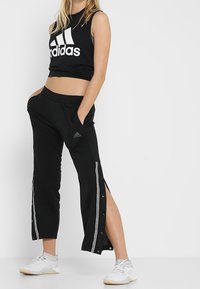 Černý crop top Adidas s bílým logem a černé kalhoty s bílými pruhy po stranách, štěrbinami na lemech a zapínáním na patentky. Bílé sportovní boty.