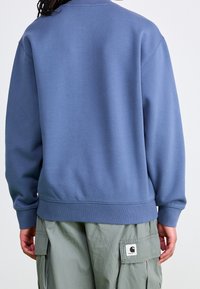Sudadera de algodón azul con cuello redondo, puños y bajo acanalados, y un logo bordado en la parte trasera. Combinada con pantalones cargo.