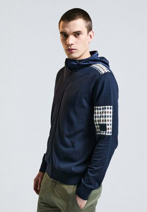 Giovane uomo che indossa una felpa con cappuccio blu navy con cerniera, con toppa a quadri sulla spalla e sulla manica, in piedi contro uno sfondo chiaro e semplice.