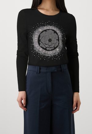 Kobieta ubrana w czarną koszulę z długim rękawem z detalicznym srebrnym kwiatowym wzorem w kształcie koła na piersi, w połączeniu z ciemnoniebieskimi spodniami z wysokim stanem.