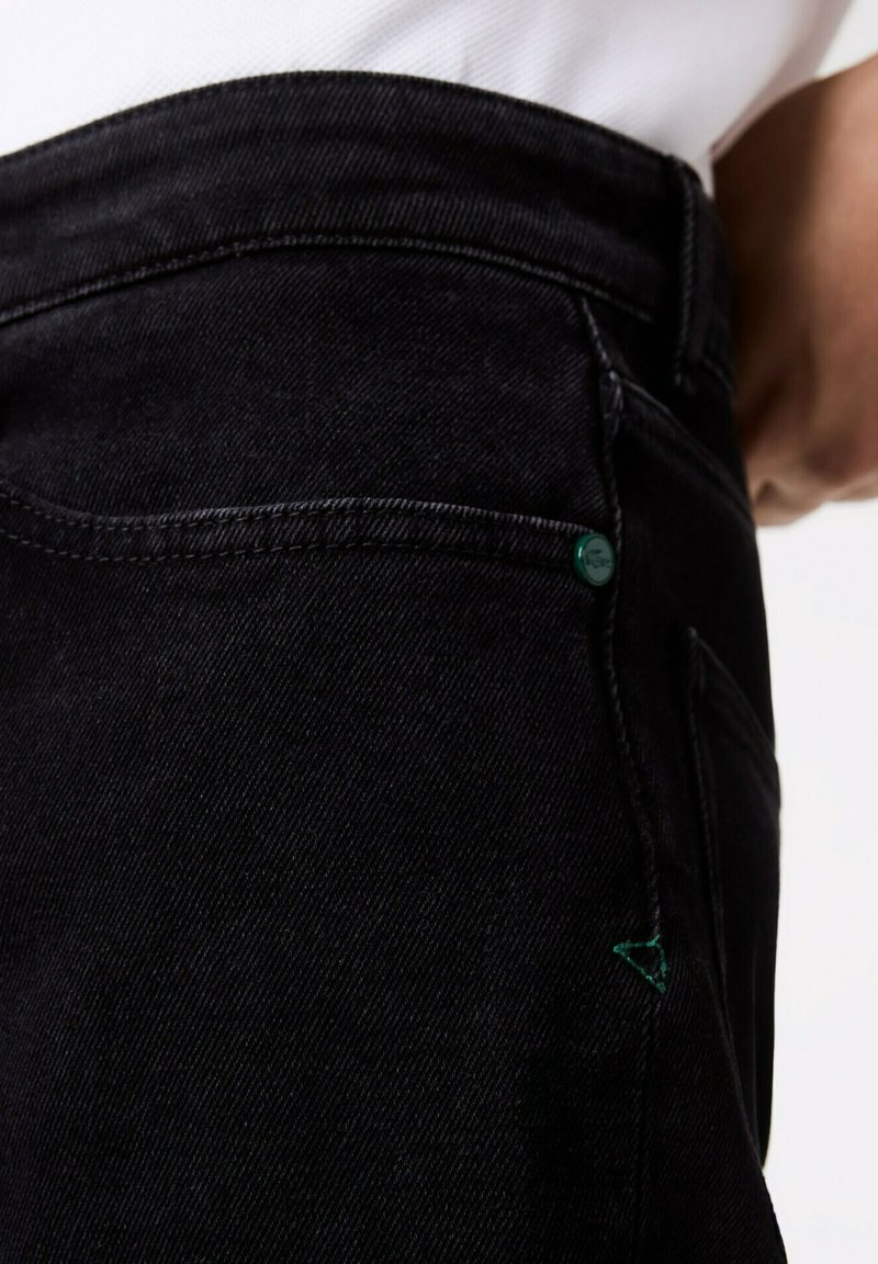 Schwarze Jeans aus Denim mit eng anliegendem Schnitt, ausgestattet mit einem grünen Akzentknopf und Nähte, die einen strukturierten Stoff und eine klare Form betonen.