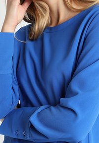 Kvinna som bär en blå blus med lång ärm och knäppta manschetter, vilande sin hand nära ansiktet.