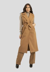 Long gilet sans manches couleur chameau avec une ceinture, doté d'un col à revers et de poches latérales, porté sur un pull ras du cou gris et associé à un pantalon beige.