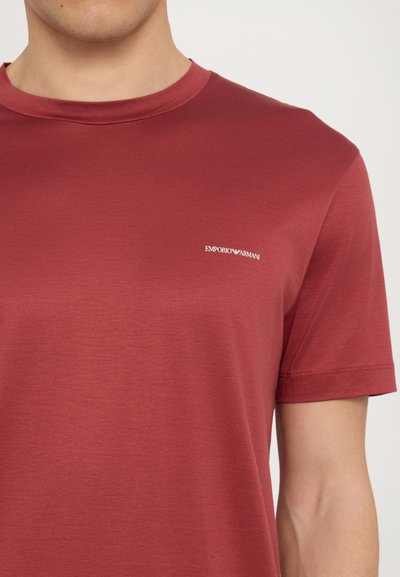 Sarkanais īsajām piedurknēm T-krekls, izgatavots no gludas auduma. Ir neliels balts Emporio Armani logo kreisajā krūšu zonā. Apaļš kakla izgriezums.