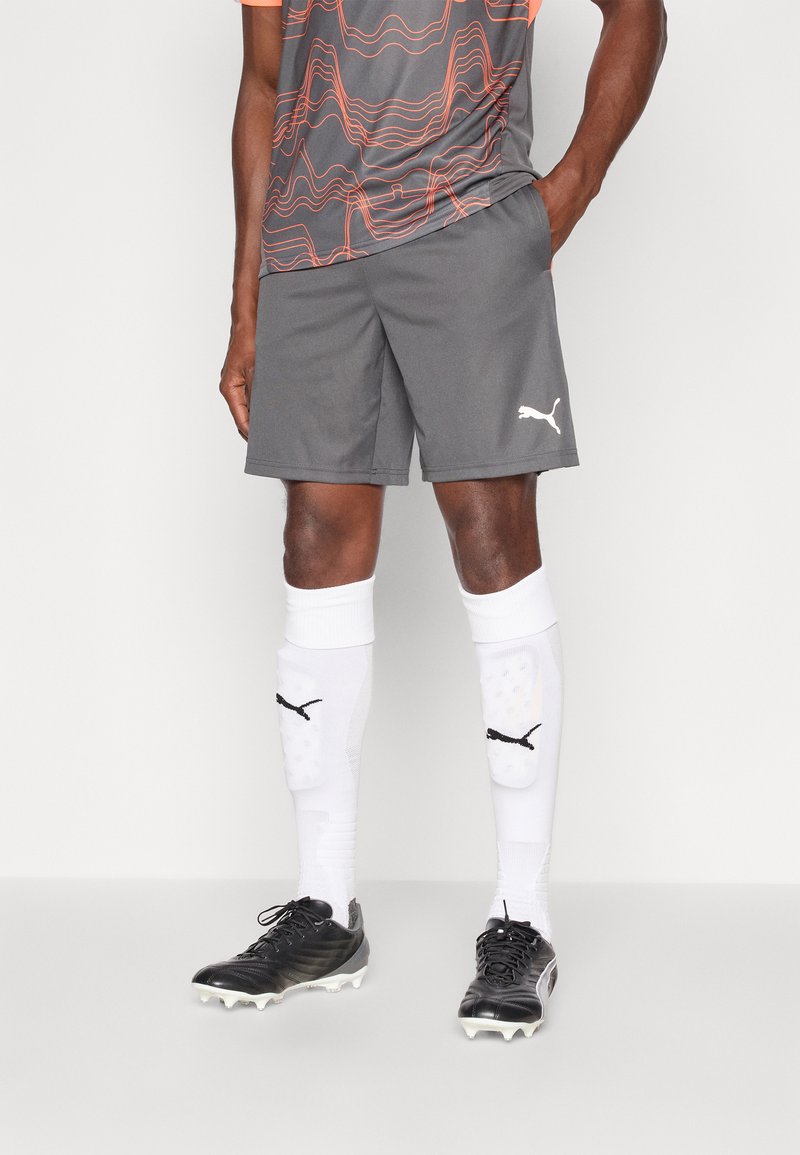 Grå sportskort, oransje mønstrete treningsskjorte, hvite sokker med sorte detaljer, og svarte fotballsko med hvite knotter.