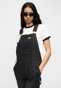 Γυναίκα με μακριά σκούρα μαλλιά που φοράει γυαλιά ηλίου cat-eye, λευκό κοντομάνικο μπλουζάκι με ρίγες και μαύρη τζιν σαλοπέτα, με τα χέρια στις τσέπες.