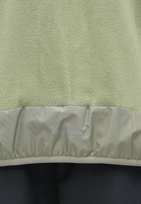 Jachetă din fleece de culoare verde deschis, cu un panou de țesătură netedă în partea de jos, având textul "ClimaWarm" în gri deschis.