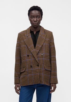 Γυναίκα με καφέ και μπλε καρό oversized διπλόκοπο σακάκι πάνω από μαύρο πουκάμισο και μπλε τζιν, στέκεται μπροστά σε απλό λευκό φόντο.