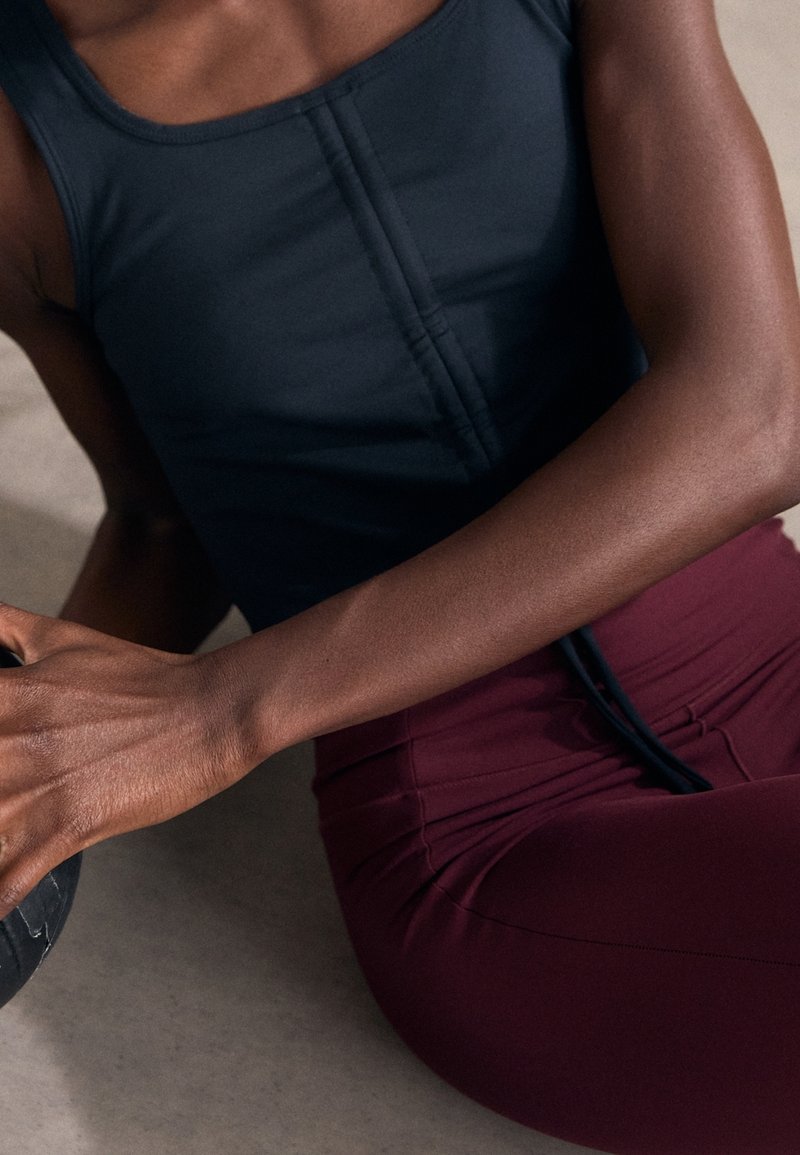 Svart sporttopp med sömmar fram, tillsammans med åtsittande bordeaux leggings. Händerna vilar på en mörk träningsboll. Slät yta.