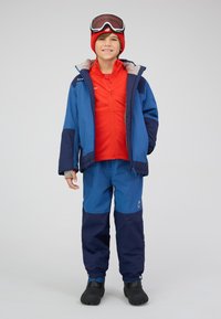 Garçon portant une veste de ski bleu et marine, un pantalon assorti, un bonnet rouge, une couche zippée rouge, des bottes de neige noires, et des lunettes de ski sur son front.