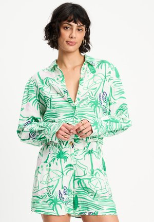 Vrouw met kort donkeren haar, gekleed in een wit hemd en een set korte broek met groene tropische print, poseert met handen samengevouwen bij haar taille.
