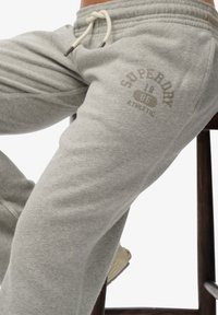 Jasnoszare spodnie dresowe z miękką teksturą, z logo „Superdry” w ciemniejszym odcieniu szarości, elastyczną talią i sznurkiem do zaciągania.