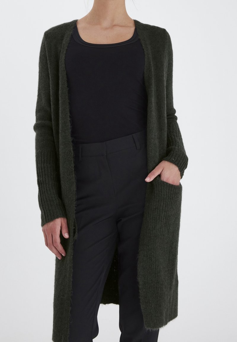 Lång, mörkgrön kofta i stickat material, med ribbade ärmar, öppen front och sidofickor. Bärs över en svart topp och byxor.