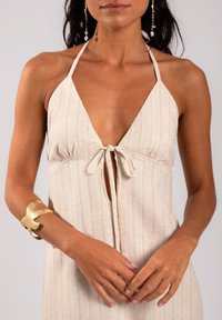 Robe halter beige clair avec des rayures verticales dorées, un détail noué à l'avant et un décolleté plongeant. Accompagnée d'un bracelet en cuir doré et de boucles d'oreilles pendantes.