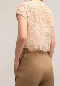 Femme portant un haut en dentelle beige transparent à manches courtes, rentré dans un pantalon camel taille haute, les mains dans les poches, vue de dos.