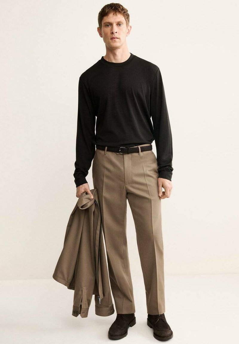 Black long-sleeve shirt, beige tailored trousers, holding a beige jacket, brown shoes, standing on a neutral background.