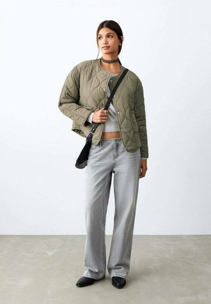 Mujer con chaqueta acolchada verde oliva, vaqueros de pierna ancha gris claro, zapatos negros puntiagudos, bolso negro de hombro y collar choker, de pie sobre un suelo gris.