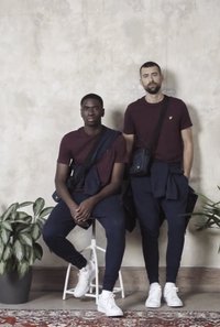 Két férfi bordó pólót és tengerészkék nadrágot visel, fehér sportcipővel párosítva. Az egyiküknek válltáskája van; mindketten mindennapi ruházatban vannak, és növényekkel pózolnak.