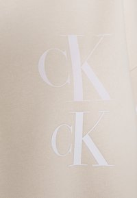 Bílý logo "ck" potištěné dvakrát na béžové texturované tkanině, uspořádané svisle, přičemž jedna sada je mírně nad druhou.