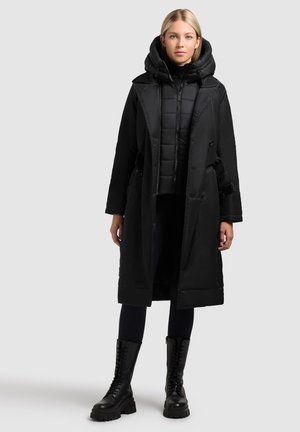 Zwarte pufferjas met een riemontwerp en een overgrote capuchon, met een gequilte textuur eronder. Gecombineerd met zwarte skinny broek en veterschoenen.