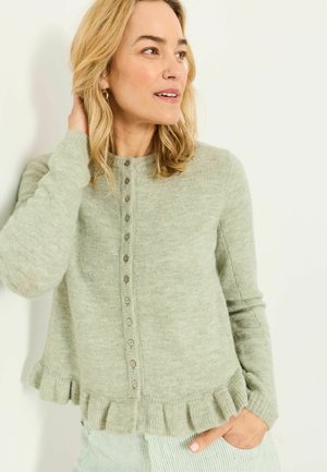 Woman wearing a light green buttoned cardigan with ruffled hem, hand in pocket, looking to her right against a plain background.